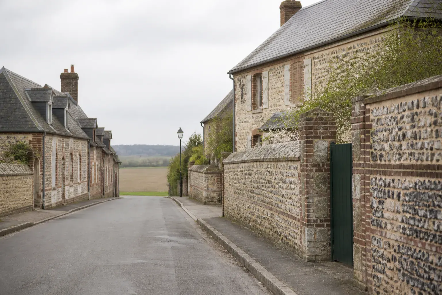 Rue d’un bourg rural du pays de Caux : maisons basses en appareillage de pierre calcaire, de brique et de silex noir taillé, murs de clôture du même appareil, portail de cour entrouvert, champs cultivés au bout de la rue